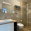 Bad mit Glas-Dusche, Toilette, Keramik-Becken und Spiegel.