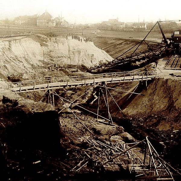 Grabenförmiger Aushub, über den eine Holzbrücke führt. Dahinter ein Braunkohlebagger und im Hintergrund Wohnhäuser.