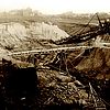 Grabenförmiger Aushub, über den eine Holzbrücke führt. Dahinter ein Braunkohlebagger und im Hintergrund Wohnhäuser.