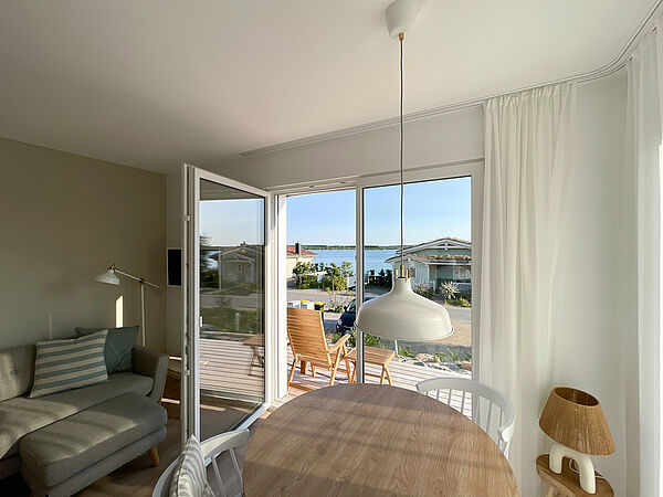 Helles Wohnzimmer mit Esstisch. Durch die geöffnete Terrassentür sieht man Holzmöbel und den Hainer See. Auf der linken Seite im Wohnzimmer steht ein Sofa und ein Fernseher hängt an der Wand. 