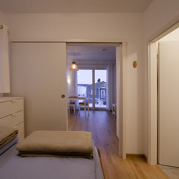 Schlafzimmer mit Doppelbett und Kommode links und einer Tür zum Bad rechts. Gegenüber ist eine geöffnete Schiebetür zum Wohnzimmer.