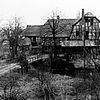 Schwarz-weiß-Foto von einem großen Fachwerkhaus am Fluss. Eine Brücke führt dort hinüber..