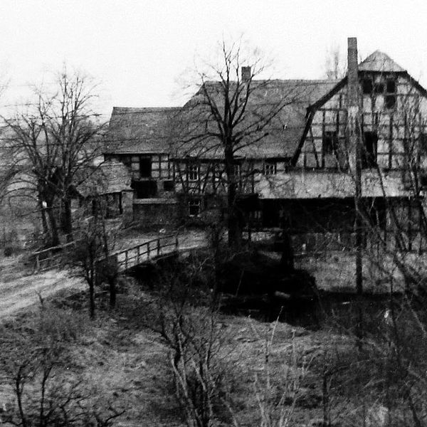 Schwarz-weiß-Foto von einem großen Fachwerkhaus am Fluss. Eine Brücke führt dort hinüber..