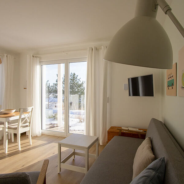 Sonniges Wohnzimmer mit großer Couch und Fernseher rechts sowie Esstisch links in der Ecke zwischen 2 bodentiefen Fenstern.