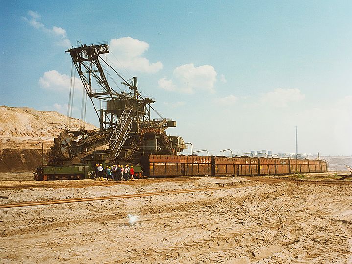 Erdlandschaft mit Braunkohlebagger. Danebne Menschen mit gelben Helmen und ein rostige Waggons. Hinter dem Zug ist ein Kraftwerk in der Ferne erkennbar.