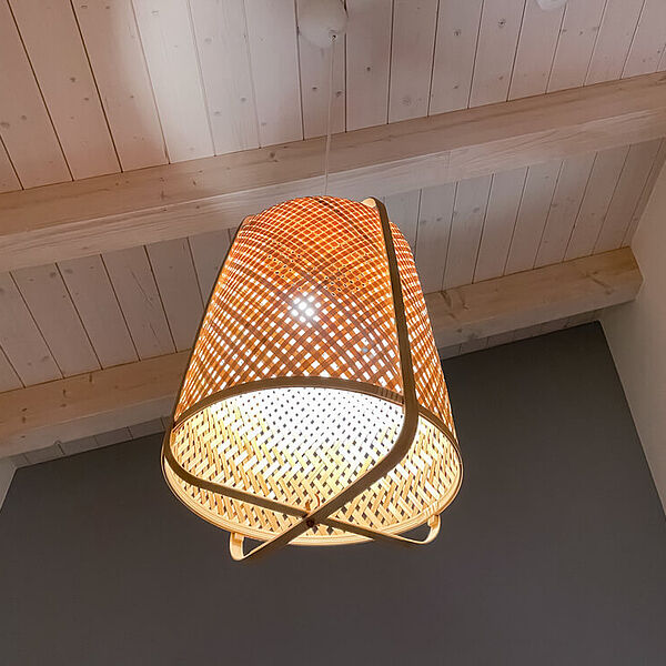 Holzdecke eines Zimmers mit leichter Dachschräge. In der Mitte hängt eine leuchtende Korblampe.