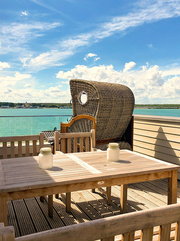 Holz-Dachterrasse mit Esstisch und Strandkorb. Im Hintergrund der grüne See unter blauem Himmel.