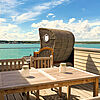 Holz-Dachterrasse mit Esstisch und Strandkorb. Im Hintergrund der grüne See unter blauem Himmel.