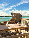 Holz-Dachterrasse mit Esstisch und Strandkorb. Im Hintergrund der grüne See unter blauem Himmel.