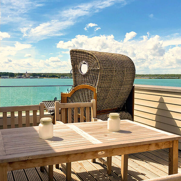 Holz-Dachterrasse mit Esstisch und Strandkorb. Im Hintergrund der grüne See unter blauem Himmel.