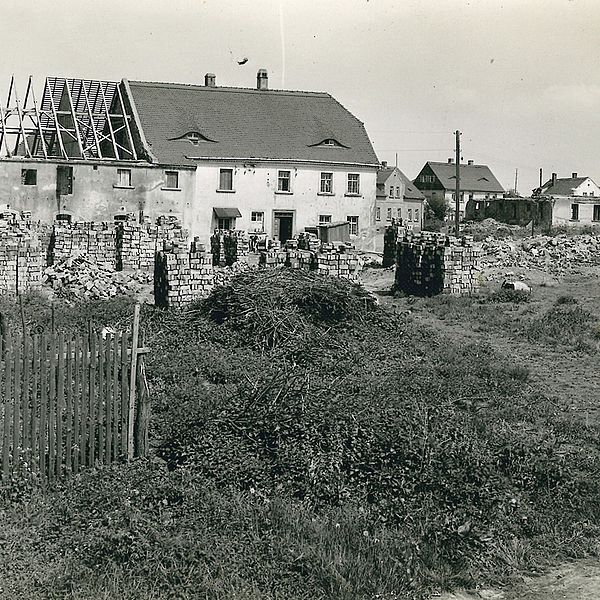 Schwarz-weiß: Haus mit abgedecktem Dach. Davor stapeln sich Ziegel- oder andere Bausteine...
