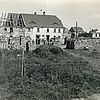 Schwarz-weiß: Haus mit abgedecktem Dach. Davor stapeln sich Ziegel- oder andere Bausteine...