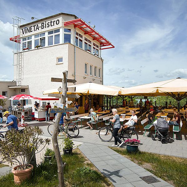 Weißer Dispatcherturm mit dem Titel "VINETA Bistro" auf der Hauswand. Davor viele Bierbänke mit Sonnenschirmen und abgestellte Fahrräder.