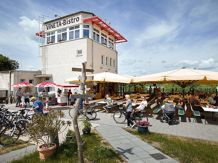 Weißer Dispatcherturm mit dem Titel "VINETA Bistro" auf der Hauswand. Davor viele Bierbänke mit Sonnenschirmen und abgestellte Fahrräder.