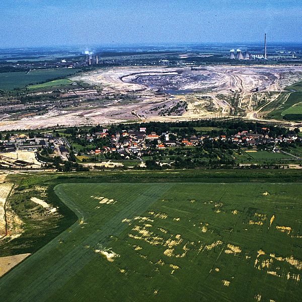 Farbige Luftaufnahme von Kahnsdorf neben grünen Feldern, direkt dahinter ein riesiges Erdloch vom Tagebau.