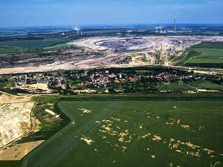 Farbige Luftaufnahme von Kahnsdorf neben grünen Feldern, direkt dahinter ein riesiges Erdloch vom Tagebau.