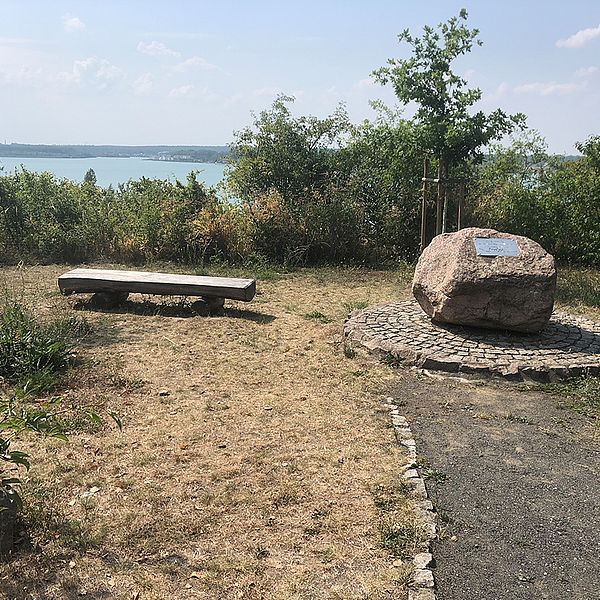 Gesteinsbrocken mit Gedenktafel, links daneben eine Bank mit Blick auf den dahinterliegenden Hainer See.