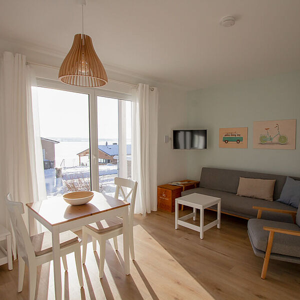 Sonniges Wohnzimmer mit Couch und Sessel rechts und einem Esstisch mit zwei Stühlen links vor einem bodentiefen Fenster. 
