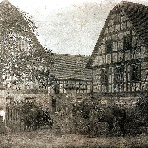 Körniges Schwarz-weiß-Foto eines Hoftores mit dahinterliegenden Fachwerkbauten. Vor dem Hoftor stehen drei Frauen mit weißen Schürzen sowie drei Männer bei Pferdefuhrwerken.