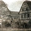 Körniges Schwarz-weiß-Foto eines Hoftores mit dahinterliegenden Fachwerkbauten. Vor dem Hoftor stehen drei Frauen mit weißen Schürzen sowie drei Männer bei Pferdefuhrwerken.