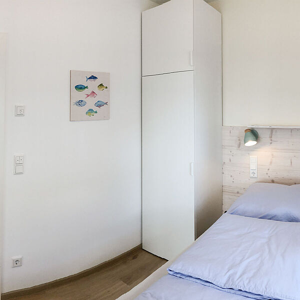 Schlafzimmer mit Kleiderschrank und Doppelbett rechts. Auf der linken Seite ist eine geöffnete Tür zu einem Badezimmer mit Waschbecken..