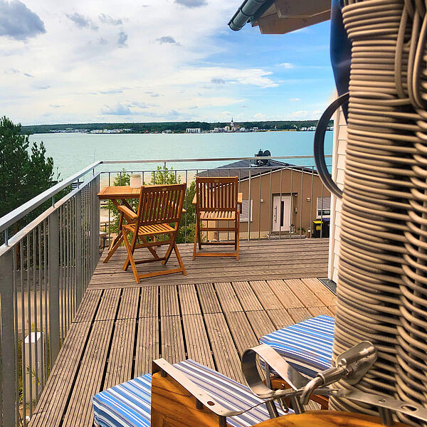 Strandkorb für zwei Personen auf dem Holzbalkon mit Blick über den Hainer See. Im Hintergrund stehen zwei Holzstühle und ein Holztisch.