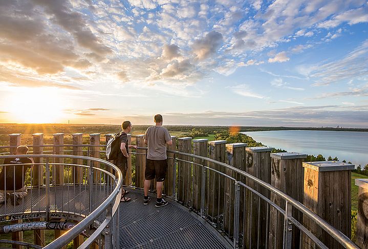 Menschen von hinten auf einem Aussichtsturm im Sommer. Auf der linken Seite ein Sonnenuntergang über der flachen Landschaft, auf der rechten Seite ein blauer See.