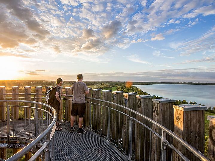 Menschen von hinten auf einem Aussichtsturm im Sommer. Auf der linken Seite ein Sonnenuntergang über der flachen Landschaft, auf der rechten Seite ein blauer See.