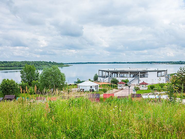 Im Vordergrund eine grüne Wiese auf einem Hügel, im Hintergrund der Schladitzer See mit Seebühne. 