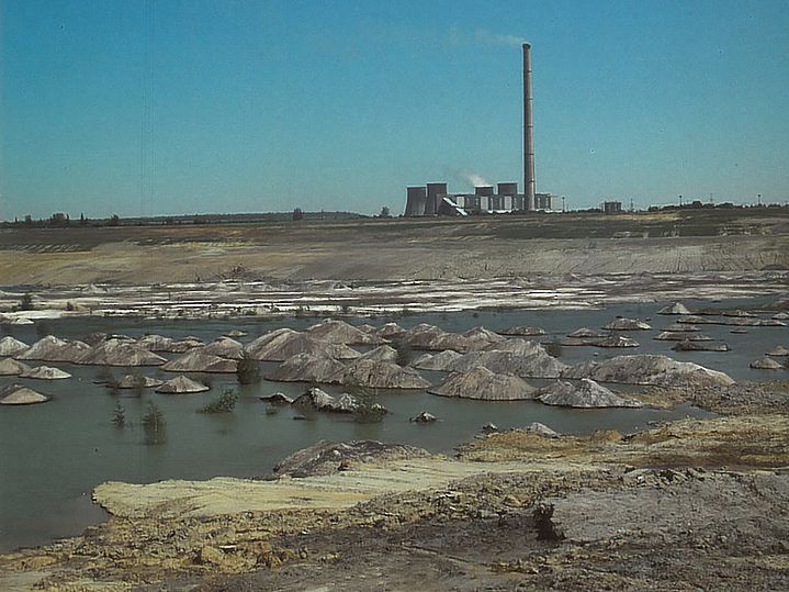 Erdige Fläche mit Wasser in Vertiefungen. Im Hintergrund ein Kraftwerk mit Schloten.