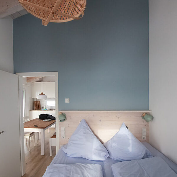 Doppelbett vor blauer Wand. An der Decke hängt eine Korblampe und links hinten führt eine Tür zum Essbereich. 