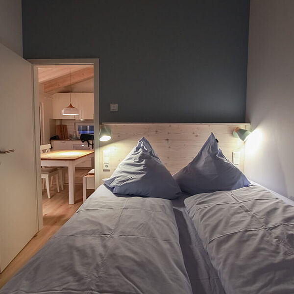 Doppelbett im verdunkelten Schlafzimmer. Links im Hintergrund eine Tür zum Essbereich..