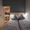 Doppelbett im verdunkelten Schlafzimmer. Links im Hintergrund eine Tür zum Essbereich..