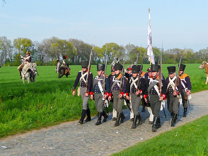 Eine Gruppe Unfiformierter marschiert auf einem Weg, im Hintergrund uniformierte Reiter.