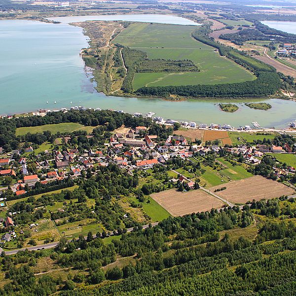 Luftaufnahme von Kahndsorf neben Feldern und Wald, dahinter direkt der blaue See.