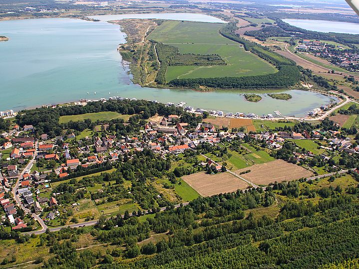 Luftaufnahme von Kahndsorf neben Feldern und Wald, dahinter direkt der blaue See.