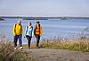 3 Menschen in Outdoor-Jacken laufen auf einem Kiesweg. Im Hintergrund ein blauer See mit Inseln im Wasser-