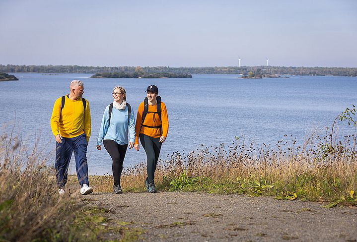 3 Menschen in Outdoor-Jacken laufen auf einem Kiesweg. Im Hintergrund ein blauer See mit Inseln im Wasser-