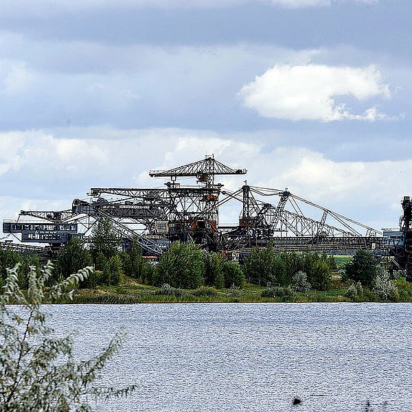 Vom Ufer aus sieht man eine Insel im Wasser, darauf sieht man die Skyline eines Tagebaubaggers.