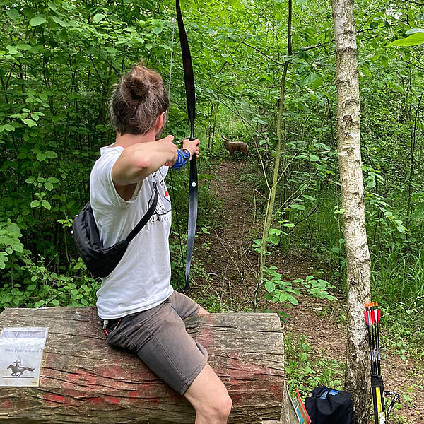 Junger Mann sitzt auf einemHolzbock und zielt mit einem Bogen auf eine Zielscheibe im Wald. Den Schützen sieht man von hinten.