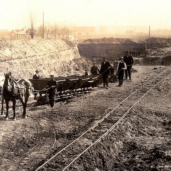 Von Pferden angetriebene Loren, die aus einem metertiefen Erdaushub fahren. Im Hintergrund Wohnhäuser.