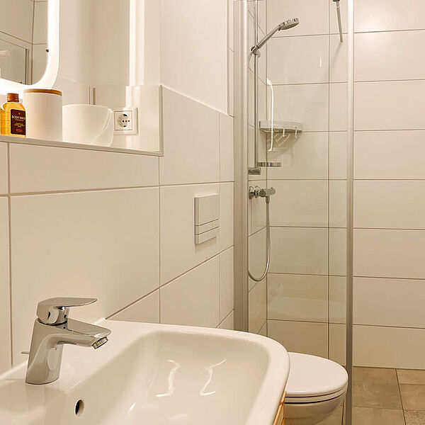  Bad mit bodentiefer Dusche, daneben eine Toilette und ein Waschbecken. Darüber eine Ablage mit Spiegel.