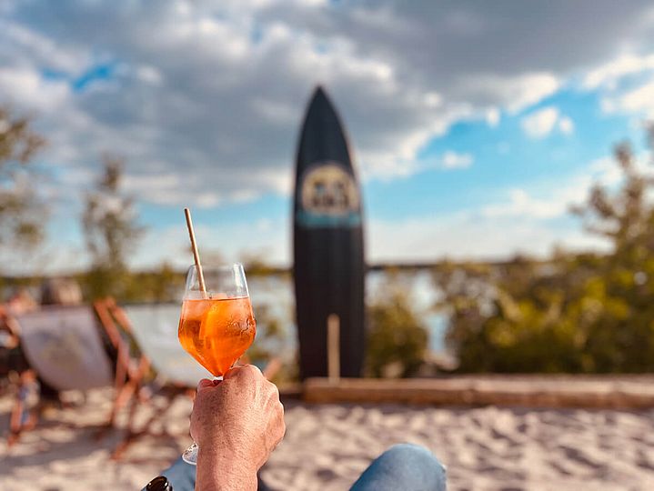 Hand mit Cocktail prostet einem Surfbrett zu