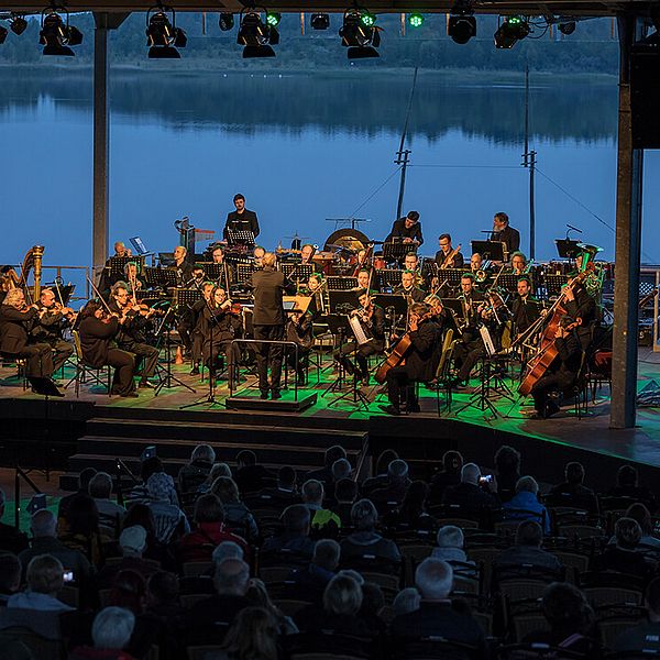 Eine Bühne mit Orchester im blauen ABendlicht, dahinter direkt der Schladitzer See.