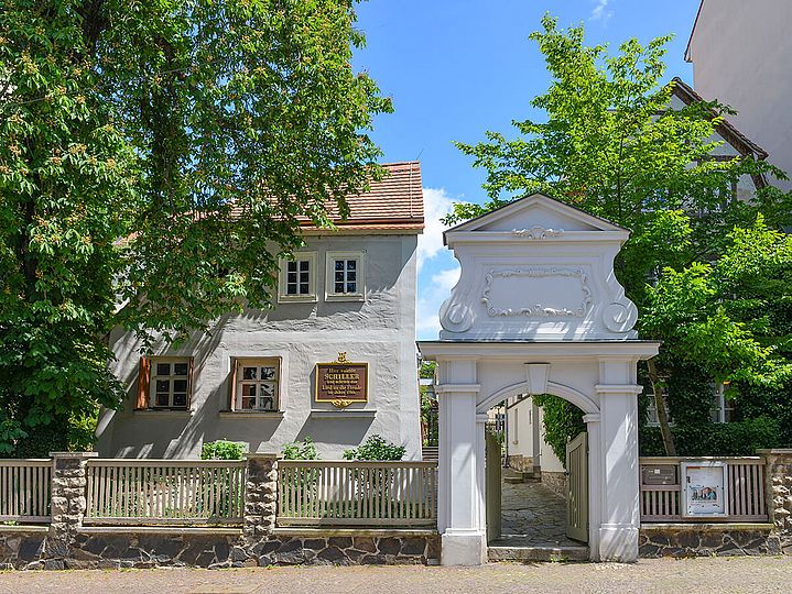 Bauernhaus mit Gedenktafel an der Wand und Garten. Ein aufwendig gestaltetes Steintor führt auf das Grundstück.