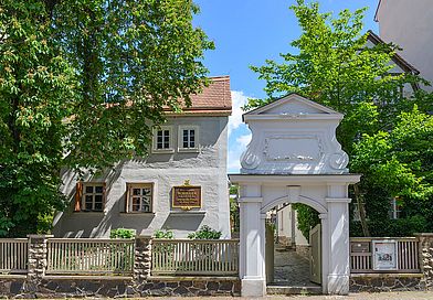 Bauernhaus mit Gedenktafel an der Wand und Garten. Ein aufwendig gestaltetes Steintor führt auf das Grundstück.