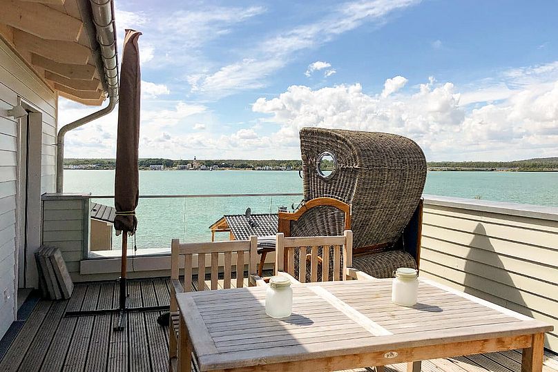 Holz-Dachterrasse mit Esstisch, Strandkorb und eingeklapptem Sonnenschirm. Im Hintergrund der grüne See.