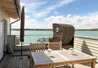 Holz-Dachterrasse mit Esstisch, Strandkorb und eingeklapptem Sonnenschirm. Im Hintergrund der grüne See.