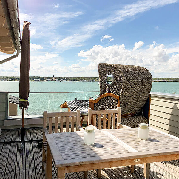 Holz-Dachterrasse mit Esstisch, Strandkorb und eingeklapptem Sonnenschirm. Im Hintergrund der grüne See.