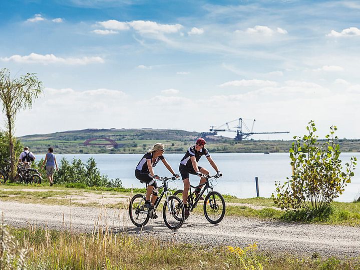 Zwei Fahrradfahrer fahren am Seeufer, im Hintergrund ein Braunkohlebagger des Bergbau-Technik-Parks